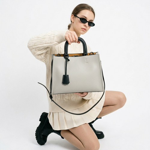 The Colette Zip Shoulder Bag in light grey with black trim, held by a model in a cream knit